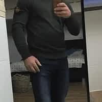 Chris Dark porn actor in grey turtle neck and jeans posing in front of a mirror. Bed in the background.