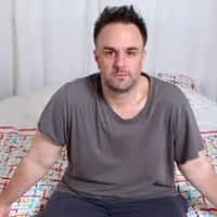 Celian Varini adult porn actor sitting down on a bed with grey shirt and blue trousers.