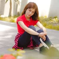 Marina Gold porn actress in red shirt and jeans, sitting on the floor behind some branches.