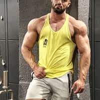 Axel Reed porn star in yellow loose gym shirt and grey shorts, lying against a wall showing his muscles.