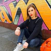 Madison MacQueen porm star sitting on the site of a street with graffiti wall behind her, in black shirt and jeans.