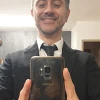 Rico Simmons pornstar in black suit, smiling white taking a citure of himself in front of a mirror.