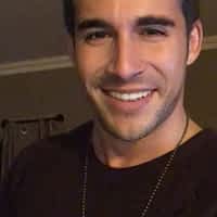 Adrian Dimas pornstar in black shirt taking a selfie and smiling. Window with beige curtain in the background.
