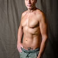 Capitano Eric pornstar shirtless wearing green trousers with one hand on his hip, posing in front of beige curtain.