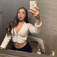 Roma Amor pornstar wearing white crop top and black trousers, taking a selfie in front of central heating.