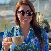 Monica Bell having a cup of coffee wearing glasses and blue floral shirt.