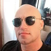 Axel Reed porno star wearing circular sun glasses and black shirt standing in front of a mirror.