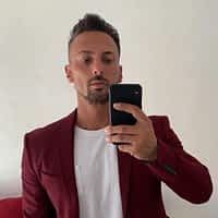 Andrew Milan porn actor in red coat and white shirt posing in front of the mirror holding his phone.