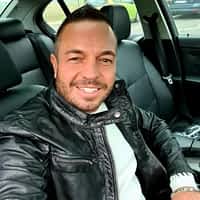 Andy Star pornstar in his car, taking a selfie wearing leather jacket and white shirt, smiling.