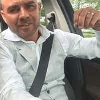 Chris Dark pornstar sitting in his car with his seatbelt on, wearing white t-shirt and grey vest, smiling.