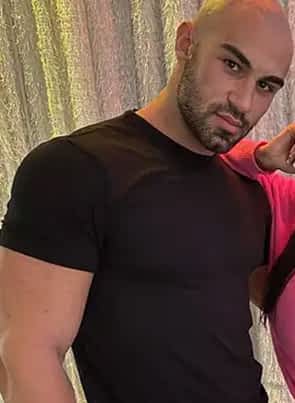 Axel Reed porn star in black shirt posing next to a woman in pink top, with silver curtain behind them.