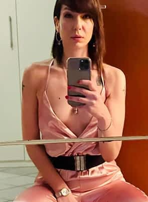 Sarah Slave pornstar sitting on a bed in her room wearing revealing pink dress and taking a picture through the mirror.