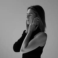 Kelly.Aleman in black and white photo, wearing a black top, touching her face with both hands. Grey background.