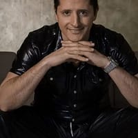 Costas Antonis pornstar in black leather clothes, sitting down on a grey couch and resting his head on his folded hands.
