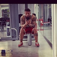 Andrew Milan porn star at teh gym in gym clothes sitting down and taking a picture with his phone from the mirror.