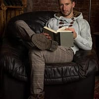 Lando Ryder porn actor sitting on a black leather couch with his legs crossed, reading a book but looking at the camera.