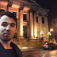 Maestro Max pornstar taking a selfie outside a large building with a car passing by.
