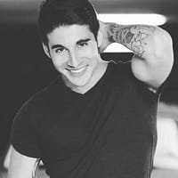 Adrian Dimas pornstar posing with black shirt and one of his hands behind his head smiling.