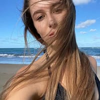 Kelly Aleman at the beach with the air blowing her hair in front of her face, she is wearing a black swimsuit.