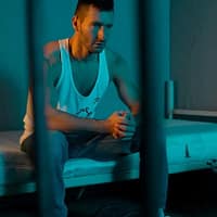 Lando Ryder porn actor behind bars wearing loose white shirt and jeans sitting on a bunk bed and clasping his hands together.