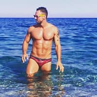 Andrew Milan male porn model, in red speedo coming out of the sea at the beach with his sun glasses.