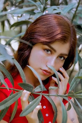 Marina Gold in red shirt touching her face with her fingers behind tree branches.